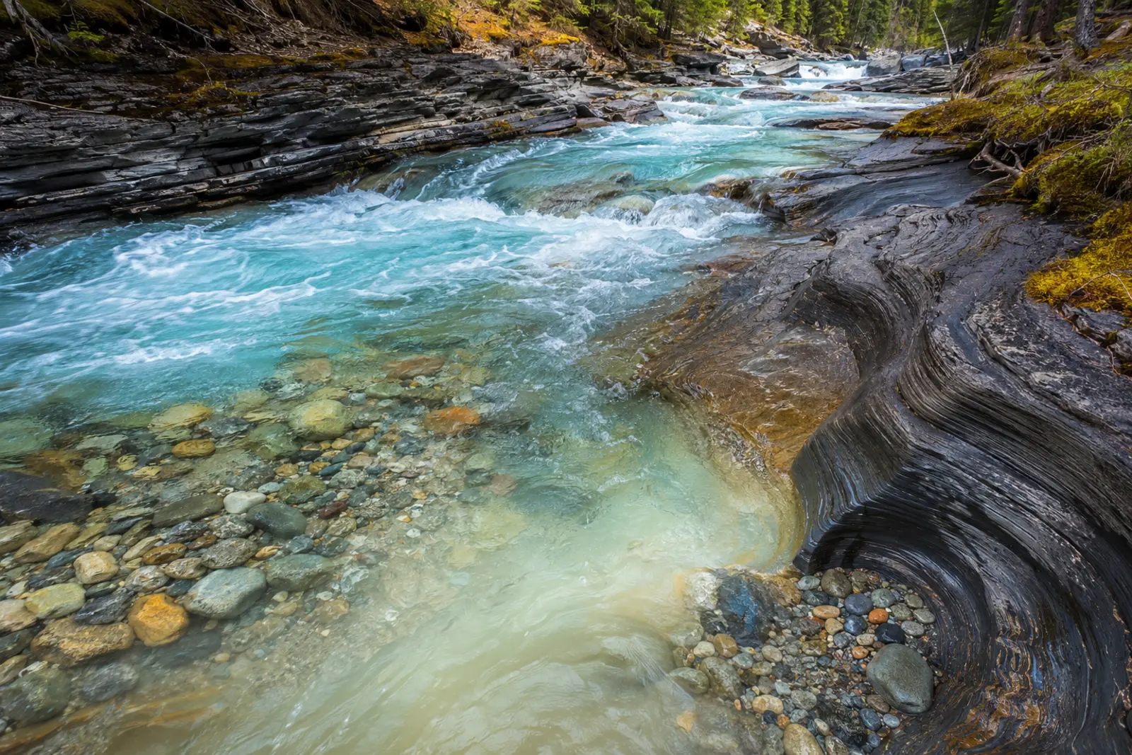 Fast turquoise stream moving around rounded pebbles and layered rock